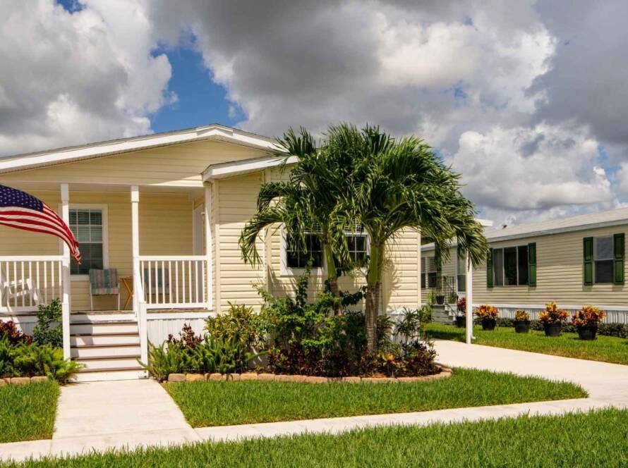 Light yellow remodeled mobile home with a front porch, American flag, and lush landscaping.